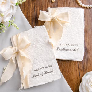 Op de afbeelding: Twee rechthoekige kaarten van gebroken wit gestructureerd papier met gescheurde randen. Elke kaart is gestrikt met een crèmekleurig lint. Eén kaart bevat de tekst "WILL YOU BE MY Maid of Honor?" en de andere "WILL YOU BE MY Bridesmaid?" in donkerbruine inkt.