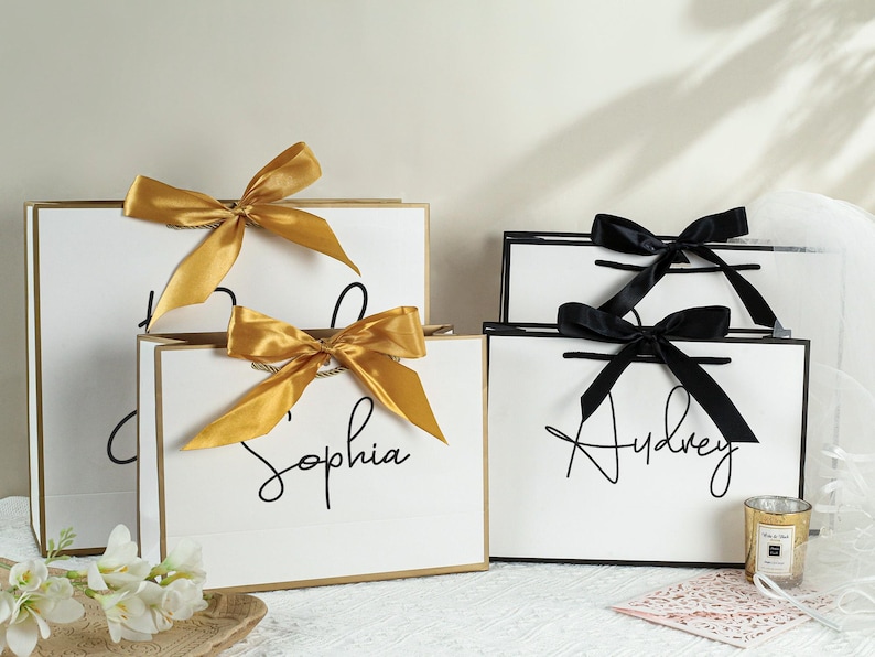 May include: Two white gift bags with gold and black ribbon bows. The bag on the left has the name "Sophia" written on it in black script. The bag on the right has the name "Audrey" written on it in black script.