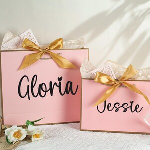 May include: Two pink gift bags with gold ribbon bows. The larger bag has the name "Gloria" written on it, and the smaller bag has the name "Jessie" written on it.