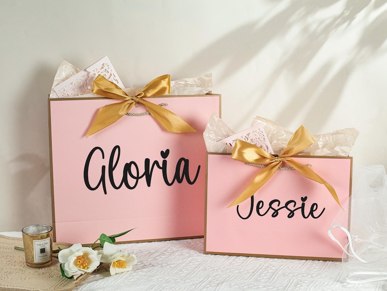 May include: Two pink gift bags with gold ribbon bows. The larger bag has the name "Gloria" written on it, and the smaller bag has the name "Jessie" written on it.