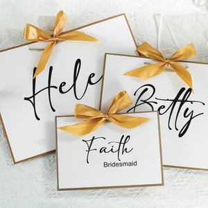 May include: Three white cards with gold ribbon bows and black calligraphy. The cards read "Helen", "Betty", and "Faith Bridesmaid".