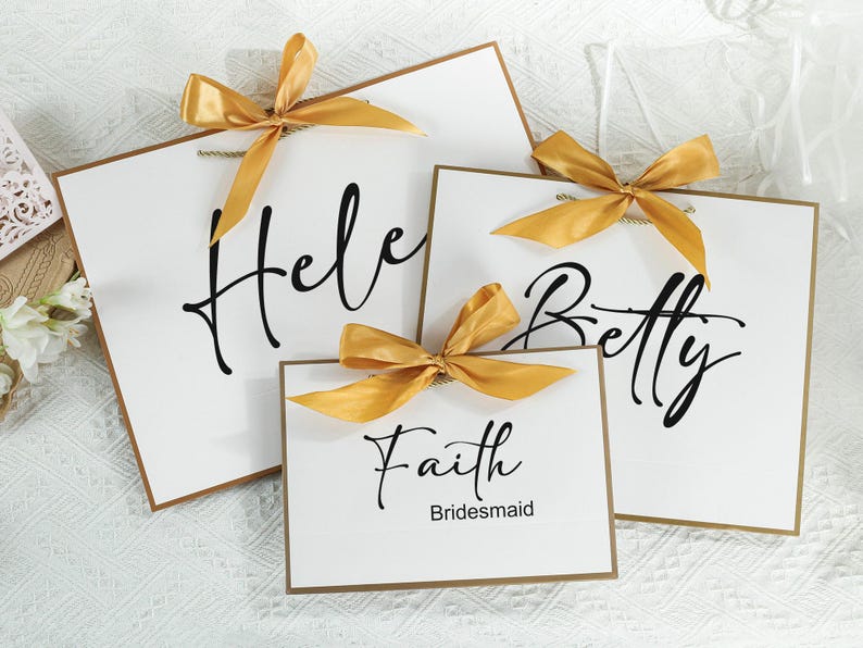 May include: Three white cards with gold ribbon bows and black calligraphy. The cards read "Helen", "Betty", and "Faith Bridesmaid".
