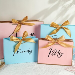 May include: Four gift bags with gold ribbon bows and personalized names. The bags are pink and blue with gold accents. The names on the bags are "Kitty", "Mandy", "Olivia", and "Sophia".