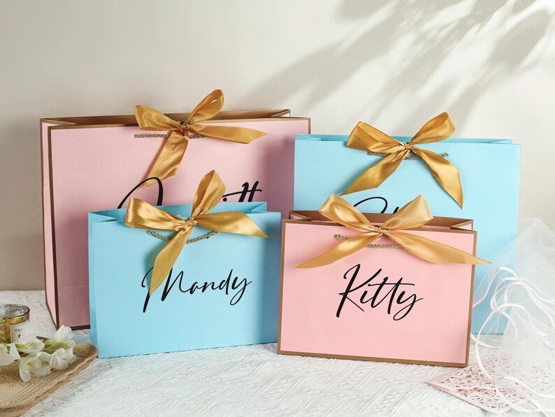 May include: Four gift bags with gold ribbon bows and personalized names. The bags are pink and blue with gold accents. The names on the bags are "Kitty", "Mandy", "Olivia", and "Sophia".