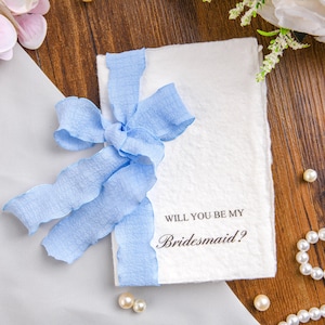Op de afbeelding: Een rechthoekige witte kaart met een getextureerde, onregelmatige rand en de in het zwart gedrukte tekst Will You Be My Bridesmaid? Een lichtblauw, gekreukt stoffen lint is in een strik rond de linkerkant van de kaart op een houten ondergrond.
