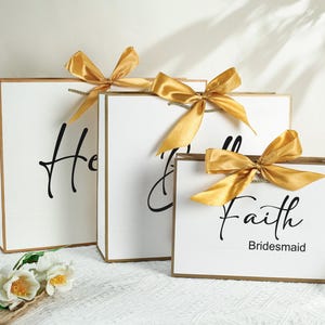 May include: Three white gift bags with gold ribbon bows. The bags are labeled "Honour", "Maid of Honour", and "Faith Bridesmaid".