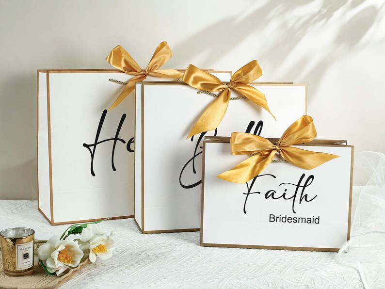 May include: Three white gift bags with gold ribbon bows. The bags are labeled "Honour", "Maid of Honour", and "Faith Bridesmaid".