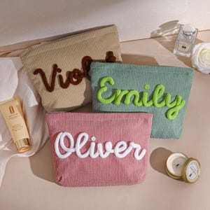 Può includere: Tre trousse per cosmetici in velluto a coste beige, verde e rosa, ciascuna con un nome ricamato in un colore a contrasto: "Violet" in marrone, "Emily" in verde e "Oliver" in bianco. Le trousse sono rettangolari con chiusura a zip.