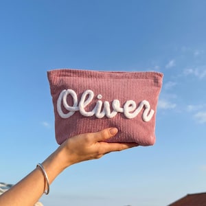 May include: A pink corduroy pouch with the name "Oliver" embroidered in white. The pouch is held up against a blue sky. A silver bracelet is visible on the person's wrist.