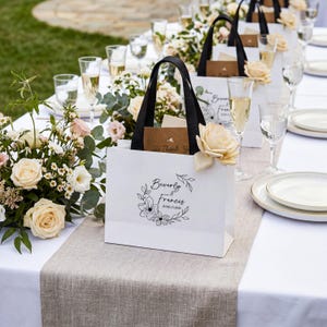 K&ouml;nnte beinhalten: Eine wei&szlig;e Geschenkt&uuml;te aus Papier mit schwarzen Henkeln, verziert mit einer floralen Kranzgrafik und dem Text Beverly & Francis JUNE 13, 2026. Die T&uuml;te ist mit einer cremefarbenen Kunstrose geschm&uuml;ckt und enth&auml;lt eine braune Karte mit dem Text Thank You.