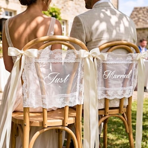 Op de afbeelding: Twee houten stoelen versierd met witte kanten hoezen met de tekst Just Married in wit schrift. Elke hoes is aan de zijkanten vastgemaakt met lange crèmekleurige satijnen linten, die strikken vormen aan de bovenkant van de rugleuningen.