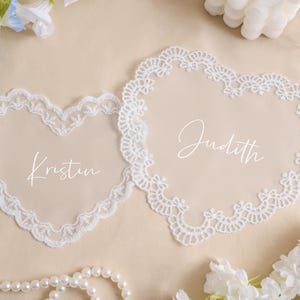 Può includere: Due sottobicchieri a forma di cuore in pizzo bianco, con i nomi "Kristin" e "Judith" in corsivo. I sottobicchieri hanno un delicato bordo in pizzo e sono su una superficie beige, con fiori e perle.