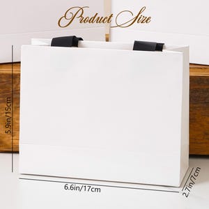 K&ouml;nnte beinhalten: Eine wei&szlig;e Geschenkt&uuml;te aus Papier mit schwarzen Bandhenkeln. Die T&uuml;te tr&auml;gt die Aufschrift Product Size und enth&auml;lt Ma&szlig;linien, die eine H&ouml;he von 15 cm, eine Breite von 17 cm und eine Tiefe von 7 cm f&uuml;r den rechteckigen Boden angeben.