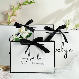 May include: Three white gift bags with black ribbon bows and personalized text. The bags are labeled "Amelia Bridesmaid", "Evelyn" and a third bag with a blank space for text.