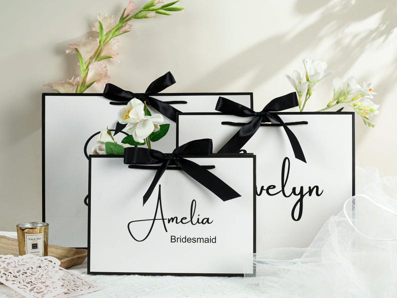 May include: Three white gift bags with black ribbon bows and personalized text. The bags are labeled "Amelia Bridesmaid", "Evelyn" and a third bag with a blank space for text.