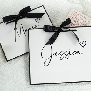 May include: Two white gift bags with black ribbon bows and black trim. The bags have the names "Maria" and "Jessica" written on them in black cursive script. Each name is accompanied by a small black heart.