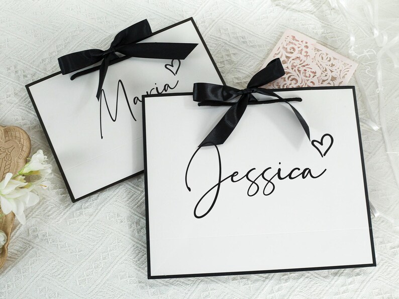 May include: Two white gift bags with black ribbon bows and black trim. The bags have the names "Maria" and "Jessica" written on them in black cursive script. Each name is accompanied by a small black heart.