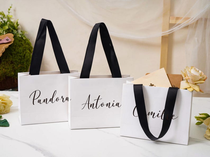 K&ouml;nnte beinhalten: Drei wei&szlig;e Geschenkt&uuml;ten aus Papier mit schwarzen Bandhenkeln. Jede T&uuml;te ist auf der Vorderseite mit einem individuellen Namen in schwarzer Schreibschrift bedruckt. Die sichtbaren Namen sind Pandora, Antonia und Camille. Rechteckige T&uuml;ten mit flachem Boden.