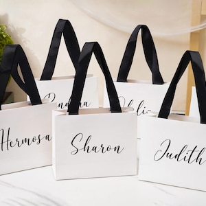 K&ouml;nnte beinhalten: Eine Ansammlung wei&szlig;er Geschenkt&uuml;ten aus Papier mit schwarzen Bandhenkeln. Jede Tasche ist mit einem in schwarzer Schreibschrift aufgedruckten Namen personalisiert, darunter die Namen Hermosa, Sharon und Judith auf der Vorderseite.