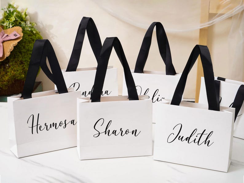 K&ouml;nnte beinhalten: Eine Ansammlung wei&szlig;er Geschenkt&uuml;ten aus Papier mit schwarzen Bandhenkeln. Jede Tasche ist mit einem in schwarzer Schreibschrift aufgedruckten Namen personalisiert, darunter die Namen Hermosa, Sharon und Judith auf der Vorderseite.