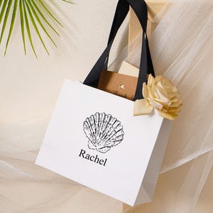 K&ouml;nnte beinhalten: Eine wei&szlig;e Geschenkt&uuml;te aus Papier mit schwarzen Bandhenkeln, die eine schwarze Strichzeichnung einer Muschel &uuml;ber dem Namen Rachel in schwarzer Schrift zeigt. Eine helle Rose und eine kleine braune Karte mit dem Text Thank You stecken darin.