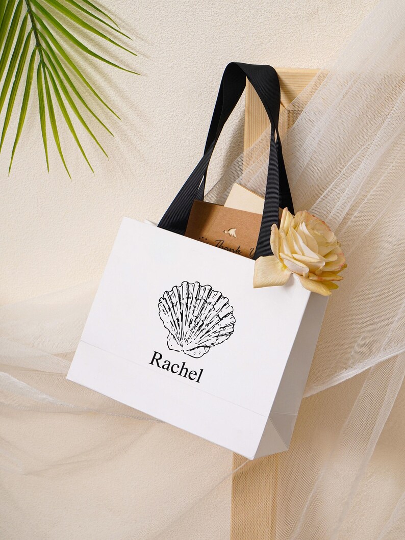 K&ouml;nnte beinhalten: Eine wei&szlig;e Geschenkt&uuml;te aus Papier mit schwarzen Bandhenkeln, die eine schwarze Strichzeichnung einer Muschel &uuml;ber dem Namen Rachel in schwarzer Schrift zeigt. Eine helle Rose und eine kleine braune Karte mit dem Text Thank You stecken darin.
