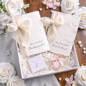 May include: Two off-white handmade paper cards with cream ribbon bows, one reading "Will you be my bridesmaid?" and the other "Will you be my maid of honor?", inside a gift box with a pearl pendant necklace and a small floral perfume bottle.
