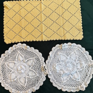 May include: A rectangular yellow crochet mat with a diamond pattern and scalloped edges, accompanied by two circular white crochet doilies featuring a central floral motif and intricate openwork borders with scalloped edges.