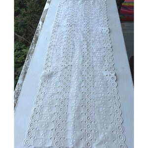 May include: A long, rectangular white table runner featuring an intricate cutwork embroidery pattern. The design consists of repeating vertical columns of circular motifs and small floral-like shapes, finished with a scalloped, embroidered edge.