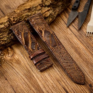 Może przedstawiać: Dwa brązowe skórzane paski do zegarków z teksturowanym wzorem, prawdopodobnie strusim. Paski są prezentowane na drewnianej powierzchni z narzędziami do obróbki skóry i szpulkami nici. Skóra ma bogaty, ciepły odcień.