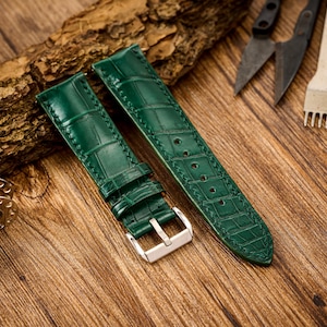 Puede incluir: Dos correas de reloj de cuero verde con detalles de costura. Las correas están dispuestas sobre una superficie de madera.