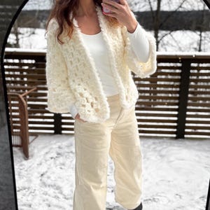 May include: Cream-colored, open-front cardigan with a textured, fluffy appearance. The cardigan is paired with a white top and light beige corduroy pants. The outfit is captured in a mirror reflection, set against a snowy outdoor backdrop.