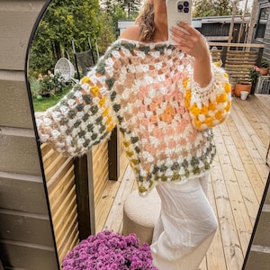 Könnte beinhalten: Ein mehrfarbiger Häkelpullover mit einem quadratischen Muster in Weiß-, Rosa-, Grün- und Gelbtönen. Der Pullover hat eine lockere Passform und einen weiten Ausschnitt.