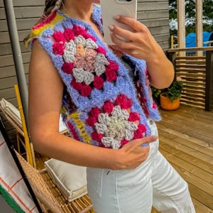 Könnte beinhalten: Eine farbenfrohe, handgefertigte Häkelweste mit Granny-Square-Design. Die Weste zeigt eine lebendige Mischung aus Farben, darunter Rosa, Blau, Gelb und Grau. Das ärmellose Kleidungsstück ist ein einzigartiges Stück tragbarer Kunst.