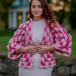 Puede incluir: Una mujer con un cárdigan rosa y rosa claro estampado. El cárdigan tiene un diseño texturizado, abierto por delante, con un patrón único. Lleva un vestido gris claro debajo. El fondo está borroso, con vegetación y un edificio visible.