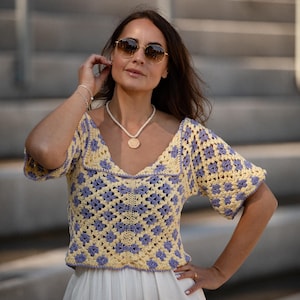 Può includere: Top all'uncinetto con un motivo floreale giallo e lavanda. Il top ha maniche corte e scollatura quadrata. La modella indossa una gonna bianca e occhiali da sole. È visibile anche una collana con un ciondolo.