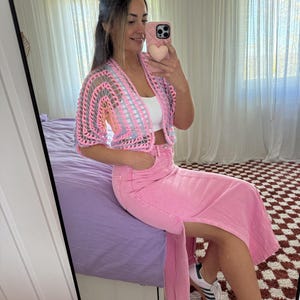 Pode incluir: Um cardigan rosa de crochê e uma saia jeans rosa a condizer. O cardigan tem um design vazado cinza e rosa. A saia tem uma fenda alta. A pessoa está usando tênis brancos com listras azuis.