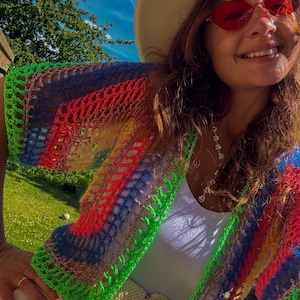 以下が含まれることがあります： 赤、青、緑、黄色のストライプ模様のカラフルなクロシェ編みカーディガン。 カーディガンは長袖で、ネオングリーンのトリムが付いています。 白いトップス、ベージュの帽子、赤いハート型のサングラスを着用しています。