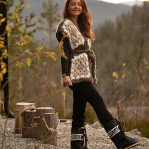 May include: A person wearing a handmade crochet vest with a geometric pattern in shades of brown, cream, and beige. They are also wearing black trousers and black winter boots. The vest is tied at the waist with a matching belt.