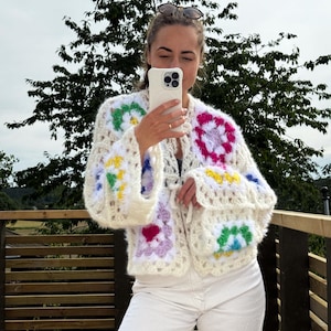 Può includere: Un cardigan bianco all'uncinetto con toppe quadrate colorate. Il cardigan ha una vestibilità ampia e una chiusura a nodo. La modella indossa pantaloni bianchi e occhiali da sole.