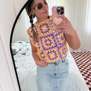 Op de afbeelding: Een kleurrijke gehaakte top in een granny square patroon, met gele, roze en paarse vierkanten. De mouwloze top heeft een gele rand. De persoon op de foto draagt een zonnebril en een lichtblauwe spijkerbroekrok.