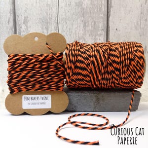 May include: Two spools of black and orange baker's twine. One spool is on a brown cardboard holder, and the other is a loose roll. The label reads "10M Bakers Twine". The Curious Cat Paperie logo is in the bottom right corner.