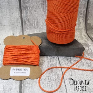 May include: Bright orange baker's twine displayed on a brown spool and a larger roll. The spool on the left has a label reading "10M Bakers Twine" and "The Curious Cat Paperie". The twine is a vivid orange colour.