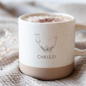 Könnte beinhalten: Weißer Keramikbecher mit gesprenkelter Oberfläche und beigefarbenem Boden. Der Becher hat ein Blumendesign mit Stethoskop und Herz, darunter steht der Name "CHRISSI". Der Becher ist mit einem Cappuccino gefüllt.