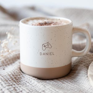 Könnte beinhalten: Ein weißer Keramikbecher mit gesprenkelter Oberfläche und einem beigefarbenen Boden. Der Becher zeigt ein kleines graues Liniengrafik-Symbol eines Videospiel-Controllers über dem Namen DANIEL in grauer Schrift. Der Becher ist mit Kaffee gefüllt.