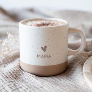 Könnte beinhalten: Weiße Keramiktasse mit einem gesprenkelten braunen Überzug. Die Tasse hat ein braunes Herz und das Wort "MAMA" darauf gedruckt. Die Tasse ist mit Kaffee gefüllt und steht auf einer beigefarbenen, strukturierten Decke.