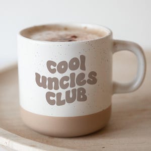 Könnte beinhalten: Eine weiße Keramiktasse mit den Worten "cool uncles CLUB" in braunen Buchstaben. Der Boden der Tasse ist einfarbig beige. Die Tasse ist mit einem Kaffeegetränk gefüllt und steht auf einem Holztablett.