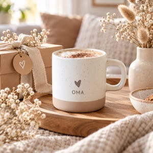 Könnte beinhalten: Ein weißer Keramikbecher mit gesprenkelter Oberfläche, hellbraunem Boden und Henkel. Der Becher zeigt ein kleines braunes Herzsymbol über dem Schriftzug OMA. Er ist mit einem schaumigen Kaffeegetränk gefüllt und steht auf einem Holztablett.