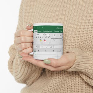 May include: White ceramic mug featuring a print of a spreadsheet interface, with green and gray elements. The mug displays the Excel interface, including the menu bar and grid. The mug is held by a person.