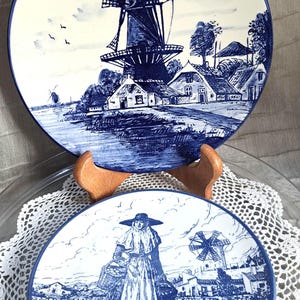 Op de afbeelding: Twee ronde keramische borden met blauw-witte illustraties. Het bovenste bord toont een windmolen en huizen bij het water. Het onderste bord toont een persoon met een hoed met brede rand die manden draagt in een veld met een windmolen en de tekst Menorca.